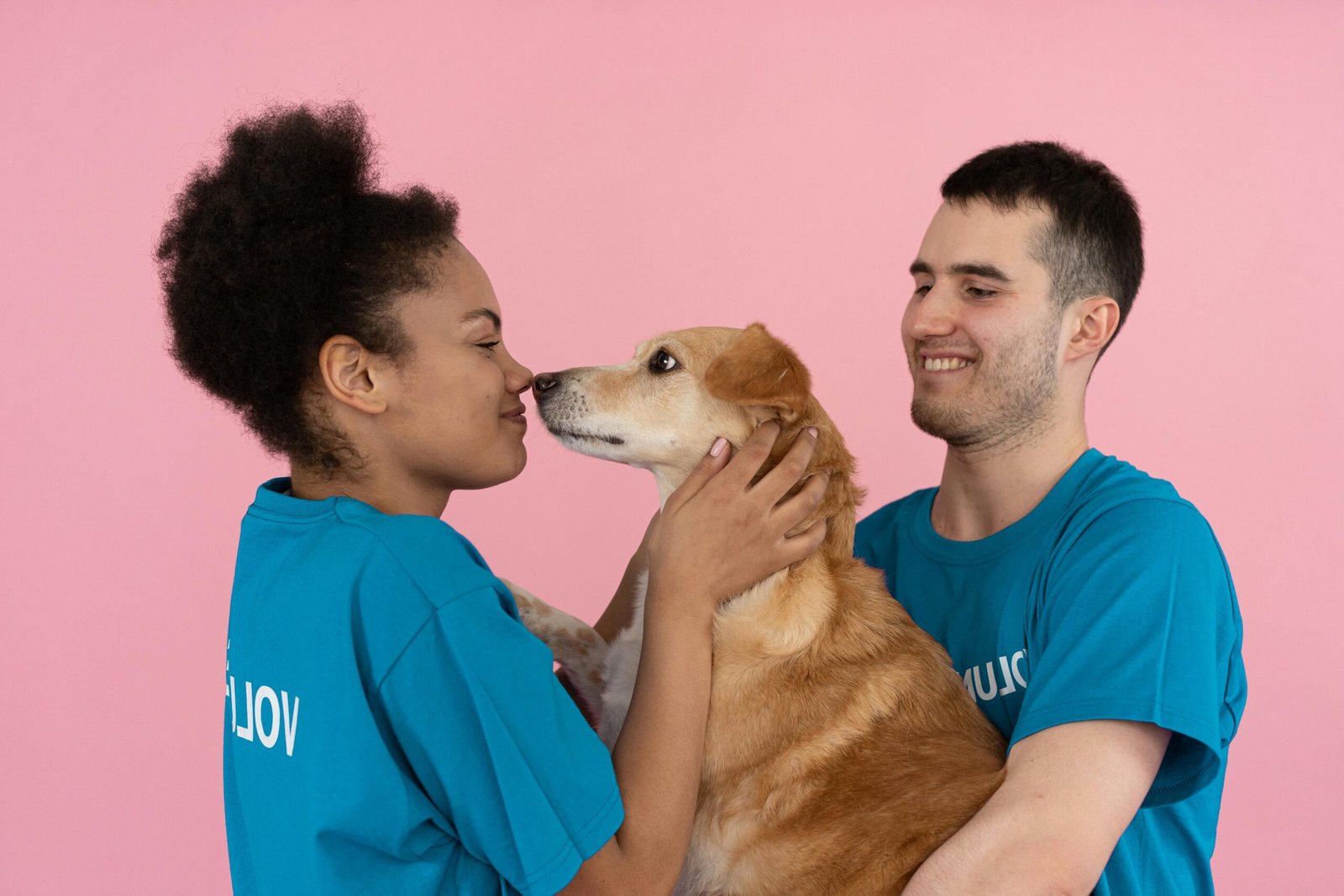 Two volunteers enjoying a happy moment with a dog against a pink background.